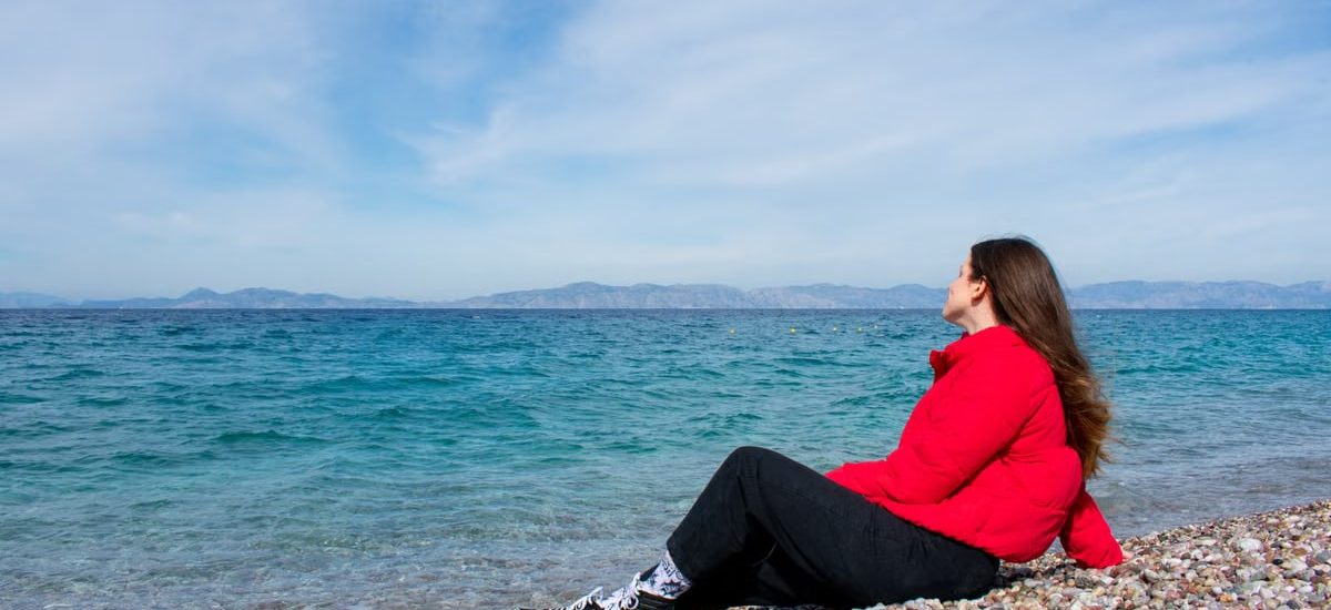 A woman in a red jacket enjoying a sunny day by the rocky beach of Greece.