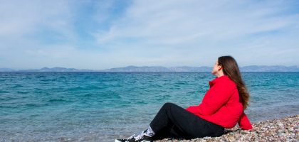 A woman in a red jacket enjoying a sunny day by the rocky beach of Greece.