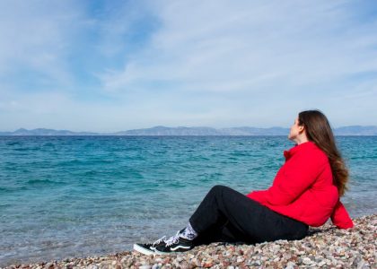 A woman in a red jacket enjoying a sunny day by the rocky beach of Greece.