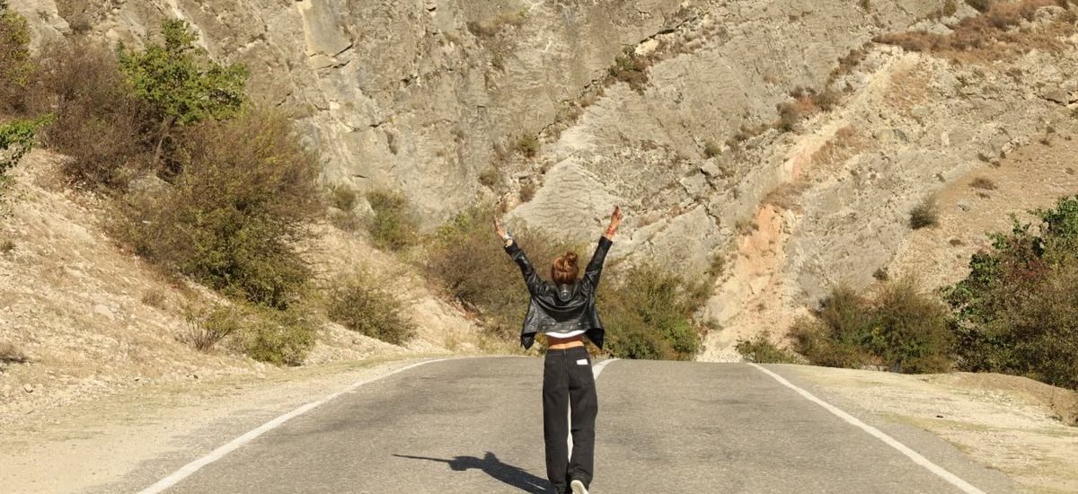 A woman in a leather jacket stands joyfully on a rural road surrounded by rocky terrain.