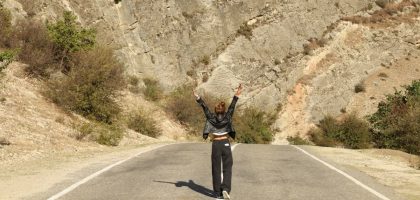 A woman in a leather jacket stands joyfully on a rural road surrounded by rocky terrain.