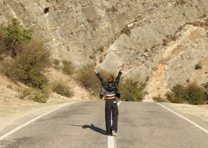 A woman in a leather jacket stands joyfully on a rural road surrounded by rocky terrain.
